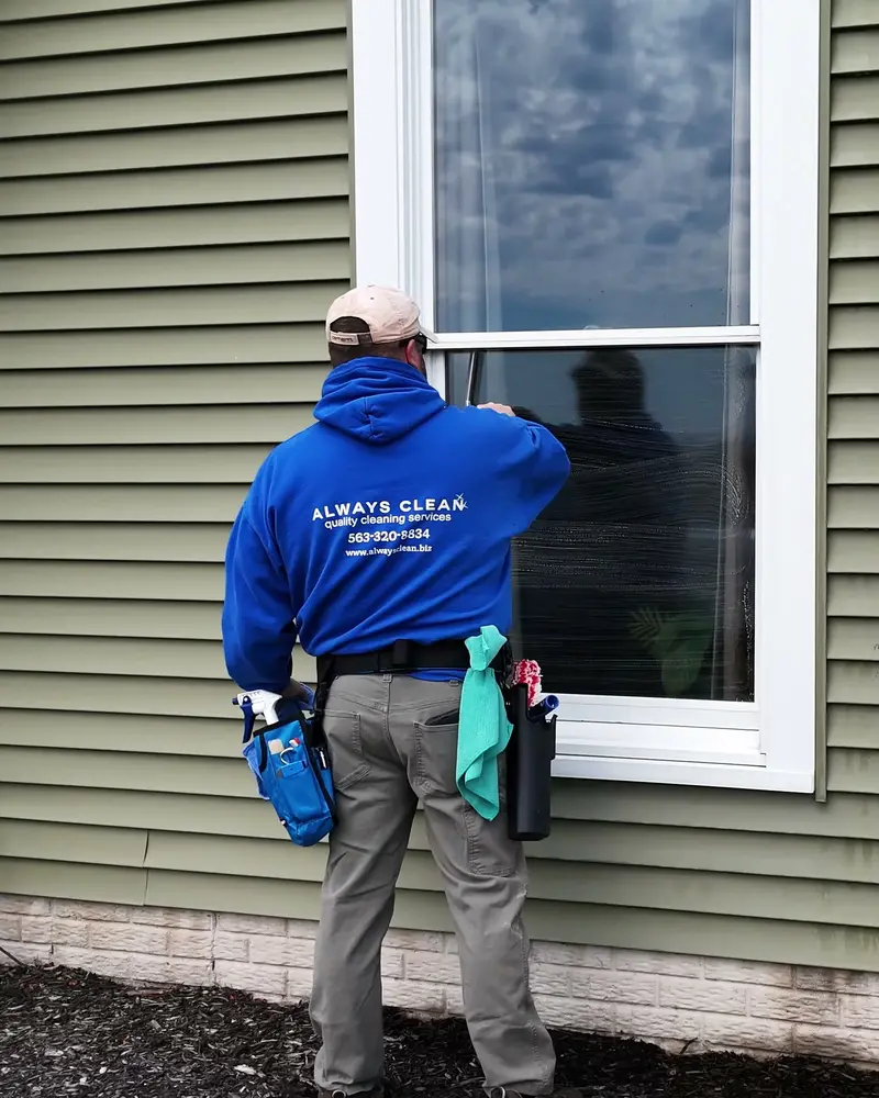 An Always Clean technician in a branded hoodie detailing a window on a Quad Cities home, squeegee and cloths on his belt