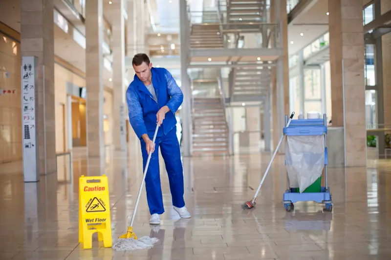 A cleaner in a blue uniform mopping the stone floor of a commercial building atrium, wet-floor sign out and janitorial cart at his side
